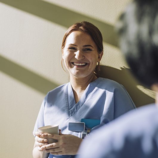 Smilende helsearbeider i blå uniform holder kaffekopp og snakker med kollega i solfylt pauseområde.